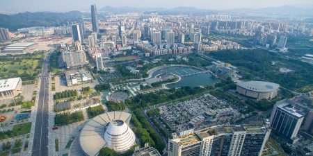 A high-angle aerial shot showcasing the cityscape of Dongguan, China, with a mix of modern skyscrapers, residential buildings, green spaces, and a large curved waterway surrounded by urban infrastructure.