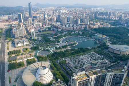 A high-angle aerial shot showcasing the cityscape of Dongguan, China, with a mix of modern skyscrapers, residential buildings, green spaces, and a large curved waterway surrounded by urban infrastructure.