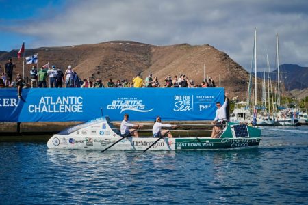East rows west team in boat pass by spectators and event banner
