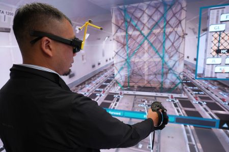 Person interacting with a 3D cargo loading simulation on a large screen, holding a controller. 