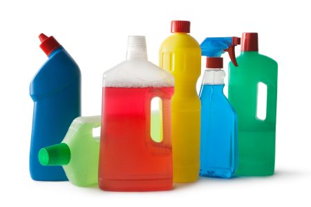 Assorted colorful plastic cleaning product bottles, including spray and liquid containers in blue, green, yellow, and clear designs, arranged together to represent household cleaning supplies