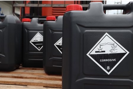 Black plastic containers with red caps labeled 'CORROSIVE' and hazard symbols showing corrosive damage to surfaces and skin, indicating Class 8 hazardous materials.